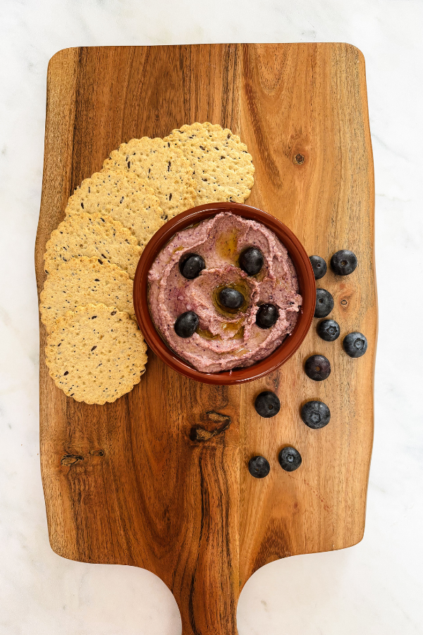 blueberry hummus served on a wooden board in a ramekin and gluten free crackers and blueberry garnish to serve